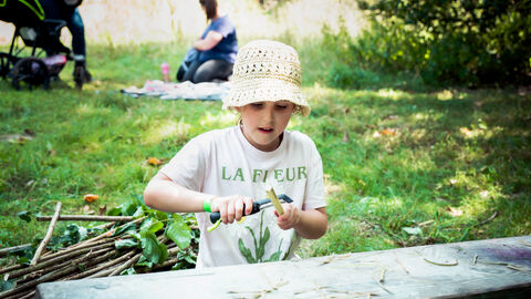 A child in the foreground wearing a floppy cream sunhat using a potato peeler to scrape away the bark of the elder twig on a summers day, whilst sat in the shade. Out of focus behind there is another family with picnic rug and pushchair. A bundle of sticks lays on the ground to one side of the child.