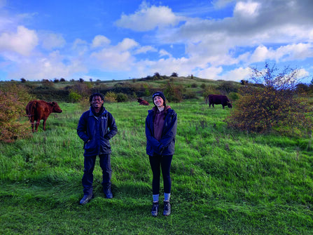 Luton trainees Aaron and Talia stand in a grassland nature reserve, with red poll cows grazing in the background